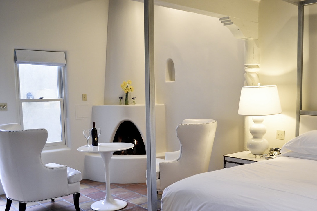 An elegant boutique hotel room with a minimalist white color scheme featuring a traditional kiva-style fireplace, likely reflecting local Southwestern or adobe architectural influences. The space includes stylish white wingback chairs, a small round table, and contemporary furnishings that blend modern design with regional elements.