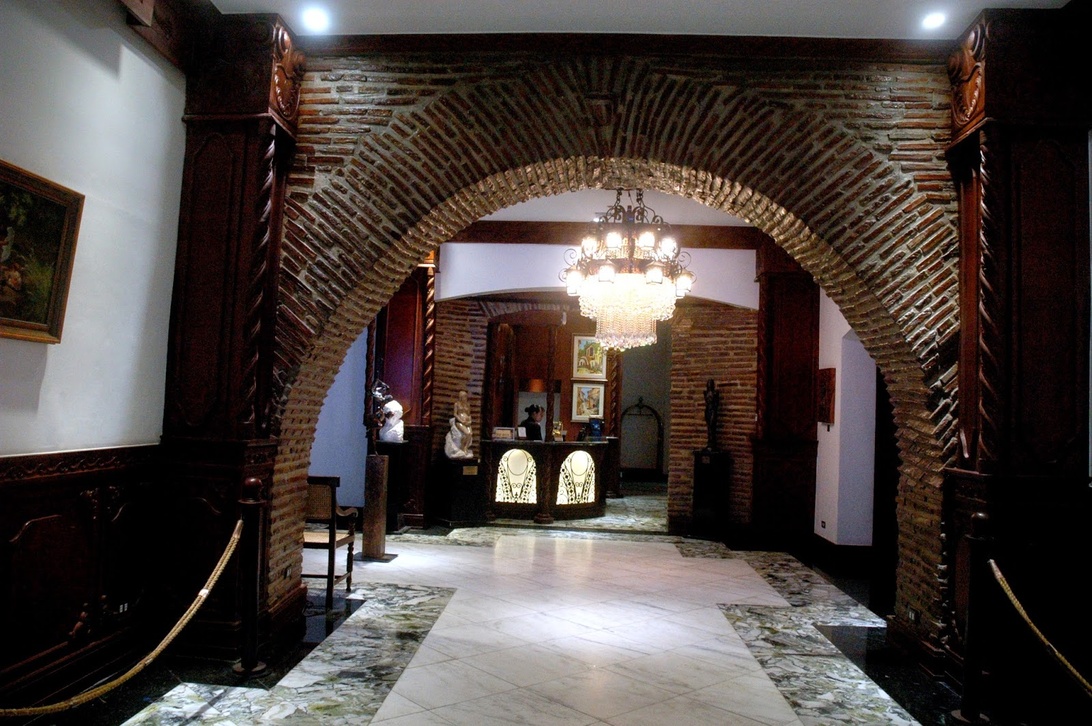An interior view of what appears to be a boutique hotel entrance or lobby featuring a large brick archway, marble flooring, wooden accents, and a reception desk visible in the background. The space has traditional architectural elements including a chandelier and some framed artwork hanging on the walls.