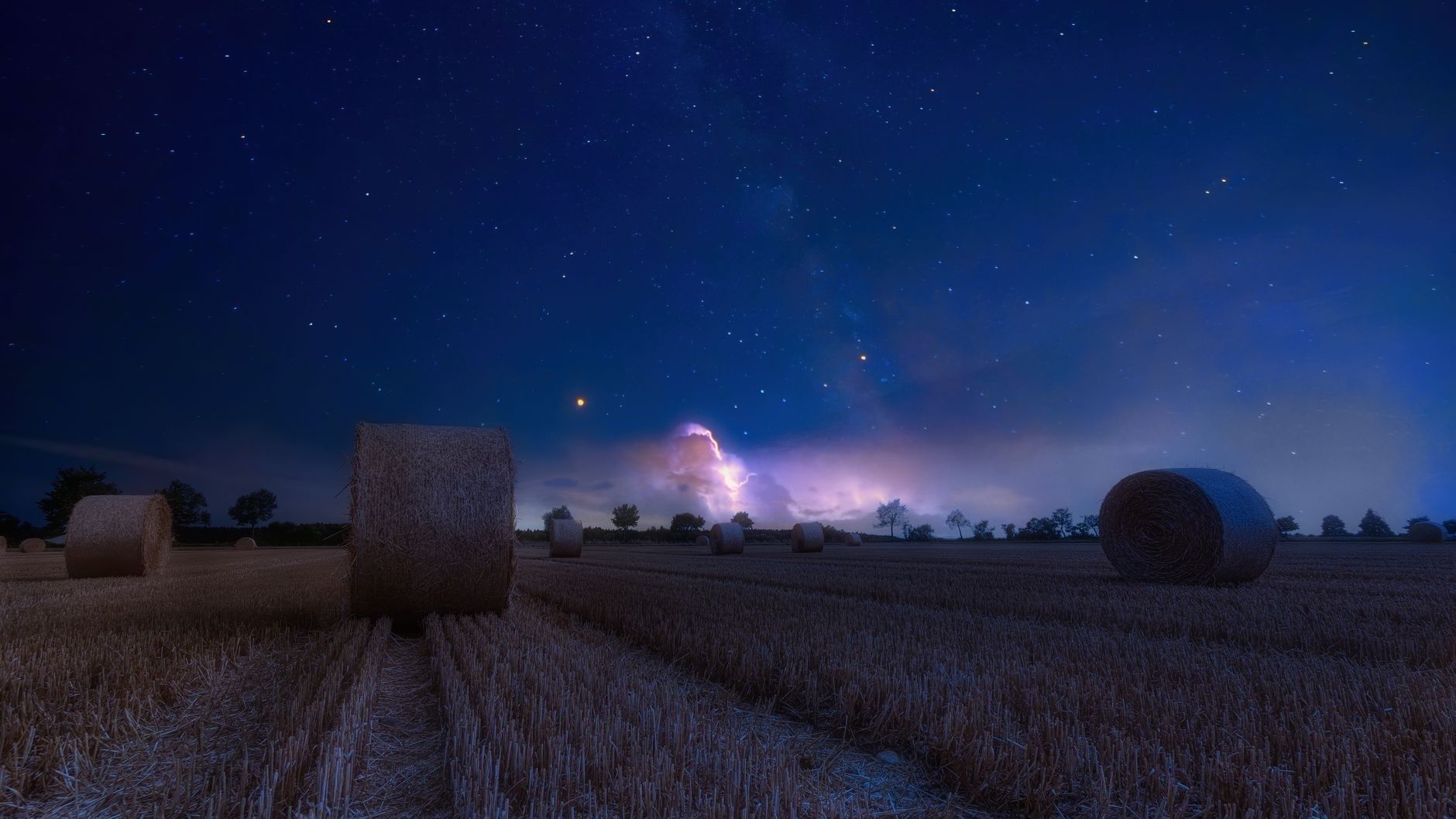 Star-filled sky and lightning over hay bales in a dark rural field